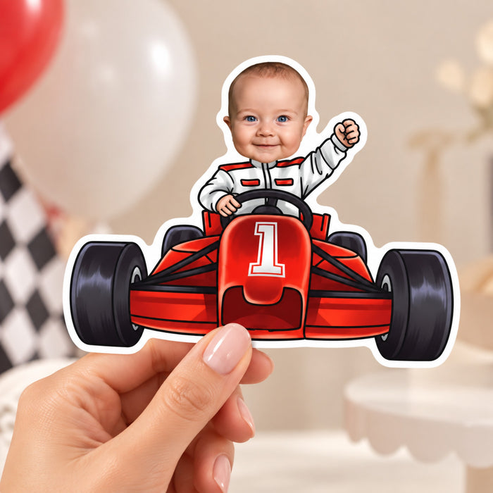 Hand holding a custom photo race car cake topper with a smiling baby driver in a white racing suit and a red number 1 race car